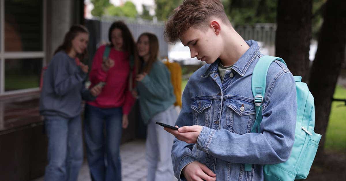 A teenage boy with a backpack looks at his phone while three other teens in the background stand together, talking and looking at him—subtle red flags that suggest they might be fake friends or not truly his friends.