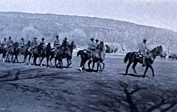 A group of people on horseback rides in a line across a grassy field with trees and hills in the background.