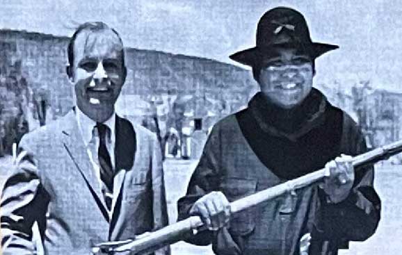 Two men standing outdoors. One is wearing a suit and tie, and the other is dressed in a hat and holding a rifle. A mountainous landscape is in the background.