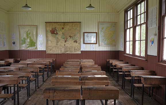 Empty vintage classroom with rows of wooden desks, world maps on walls, and large windows allowing natural light.
