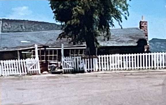 A rustic house with a white picket fence and chimney, set against a backdrop of trees and hills under a clear blue sky.