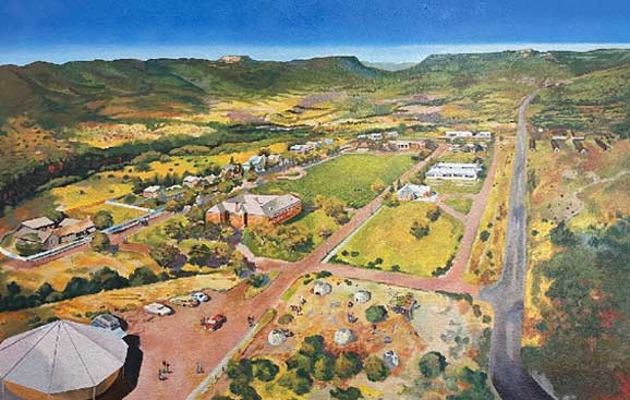 Aerial view of a rural town with buildings, roads, and green fields surrounded by hills under a clear blue sky.
