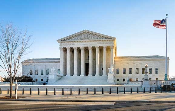The United States Supreme Court building with its neoclassical facade under a clear blue sky, an American flag flying nearby.