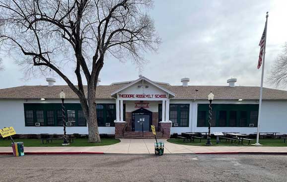 A one-story school building with "Theodore Roosevelt School" written above the entrance. An American flag is on the right, and barren trees are in the background.