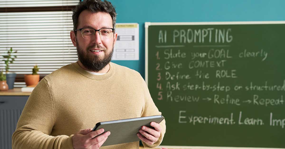 Man holding a tablet stands in front of a chalkboard with "AI Prompting" instructions written on it, highlighting the use of AI tools in a classroom setting.