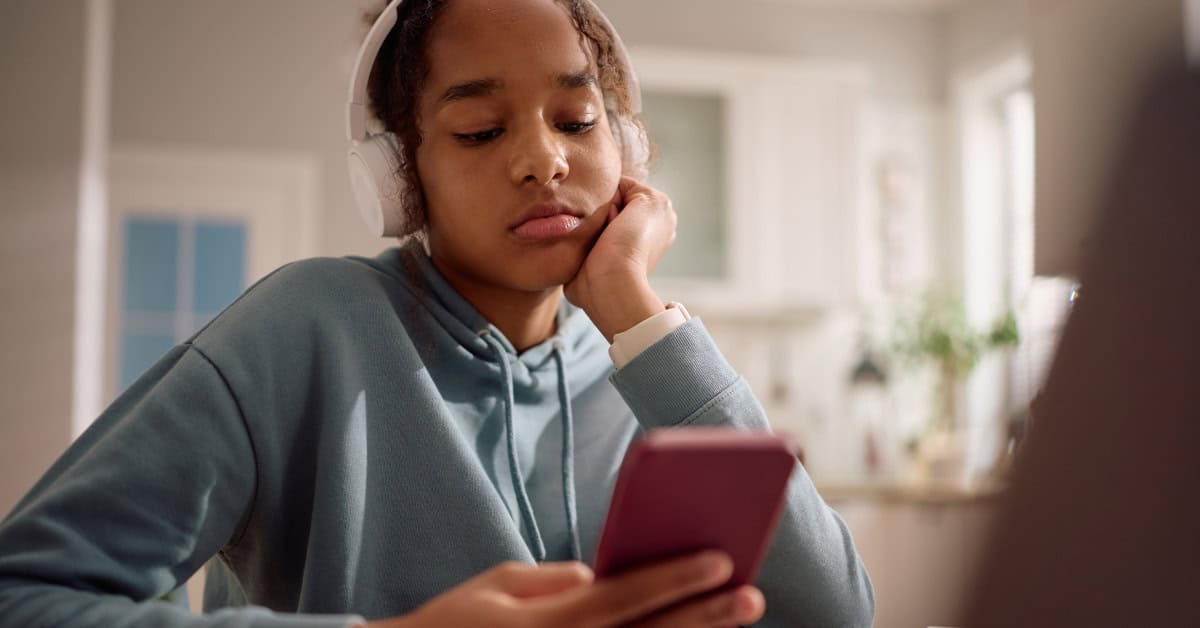 A young person wearing headphones and a blue hoodie sits at a table in a well-lit room, looking at their smartphone with a bored expression, perhaps scrolling through citizenship resources.