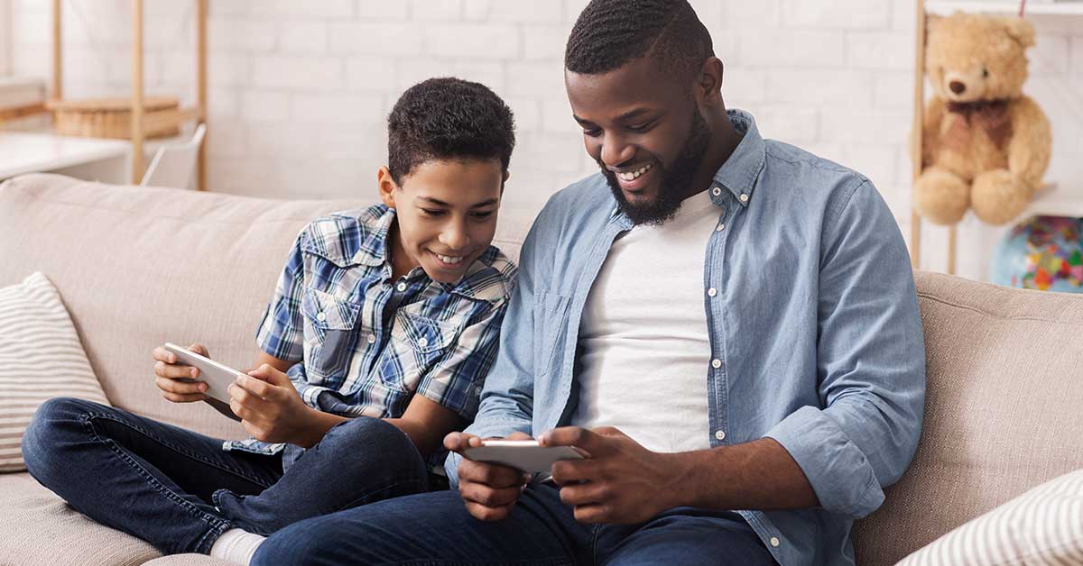 Two people sit on a couch, smiling as they play online games on handheld devices in a cozy living room, with a teddy bear in the background.