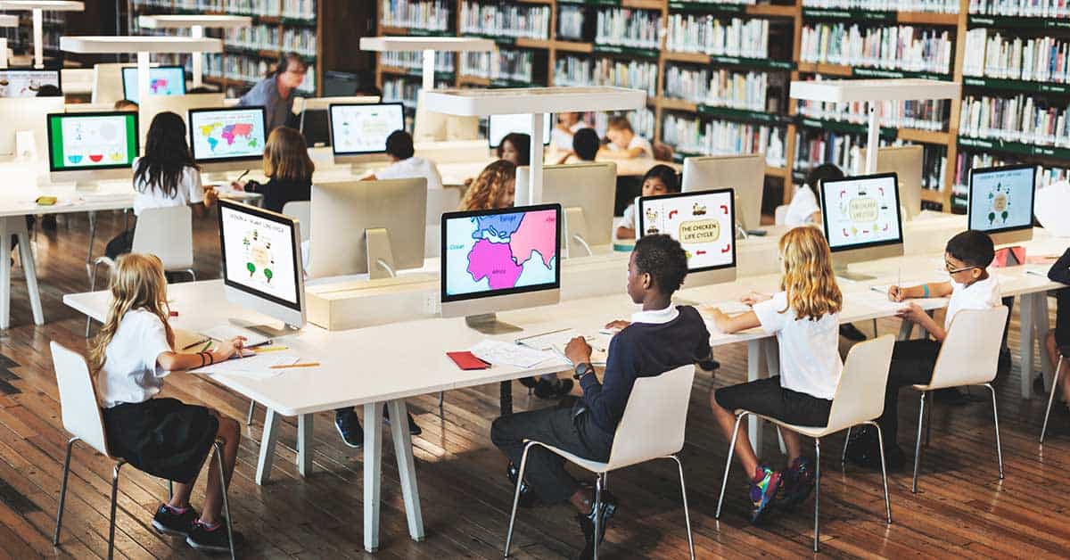 Students sit at desks using desktop computers in a library, with bookshelves in the background and educational content on the monitors, including information about deepfake technology.
