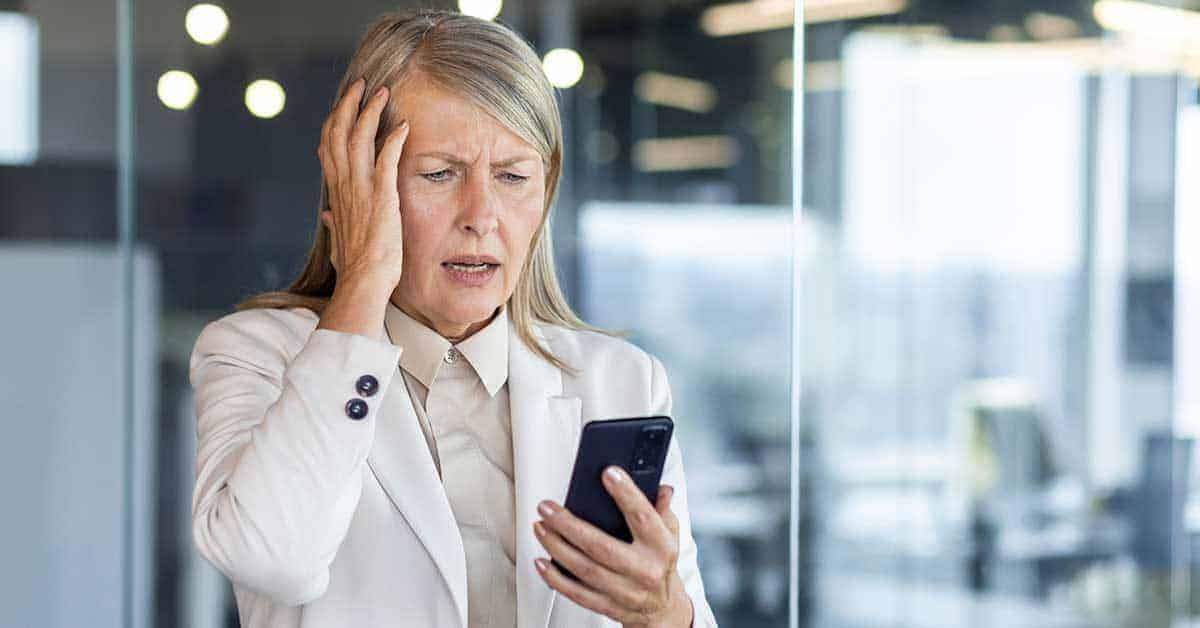 An older woman wearing a light blazer looks at her smartphone with a concerned expression, holding one hand to the side of her head in an office setting.