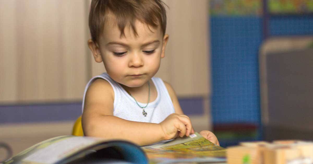 A young child wearing a sleeveless shirt sits at a table, looking at and turning the page of an open book.