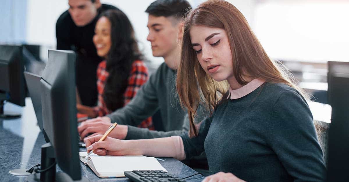 Four students sit at a row of computers. The student in the foreground writes in a notebook while using a keyboard, possibly researching ways to spot misinformation. The others focus intently on their screens.