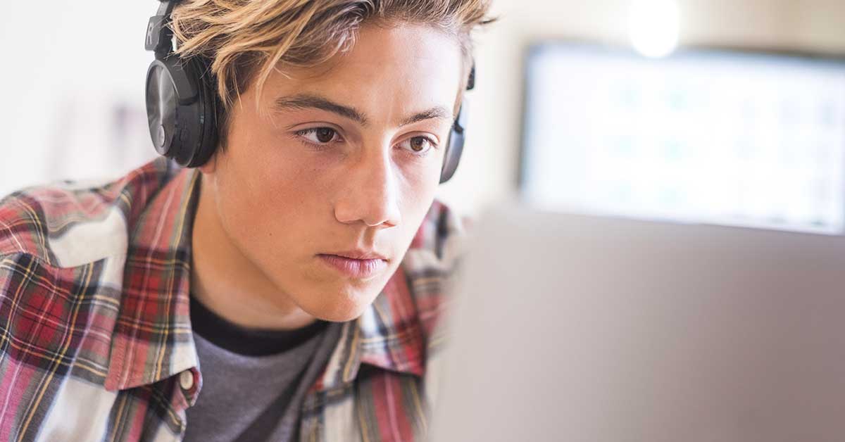 A young person wearing headphones and a plaid shirt looks intently at a laptop screen, fully immersed in a digital experience.