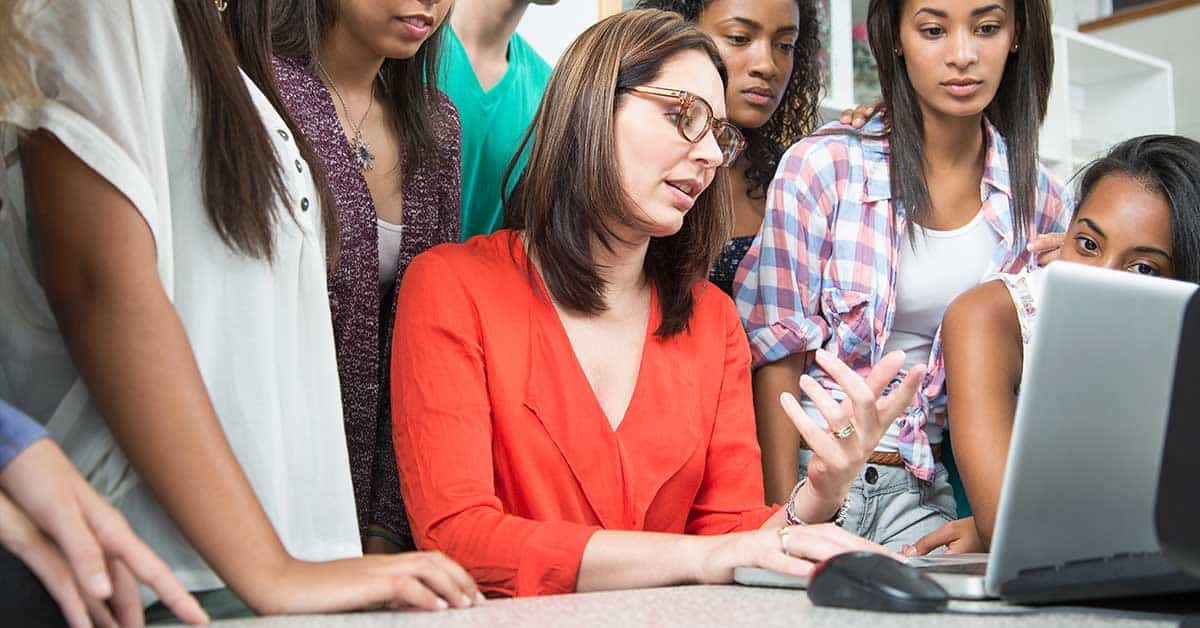A woman in glasses is seated at a desk, talking and gesturing while several young people stand around her, all looking at a laptop screen as they discuss the topic of exploitation.