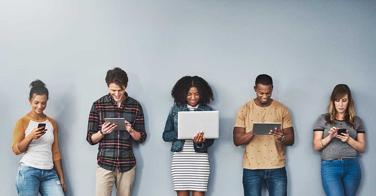 Five people stand against a gray wall, each using a digital device such as a smartphone, tablet, or laptop, fully focused on their screens.