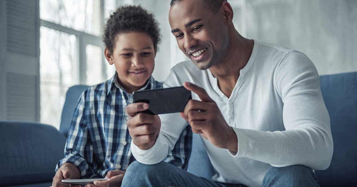A man and a boy sit on a couch, smiling and looking at a smartphone together; the boy also holds a tablet, enjoying some gaming.