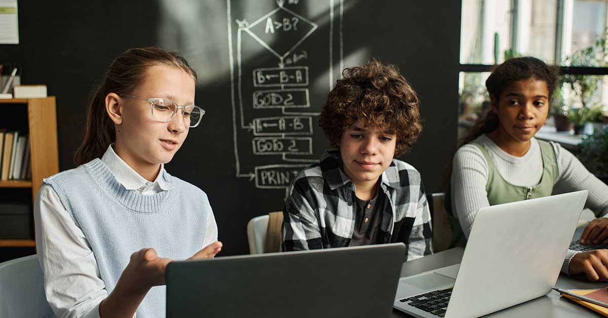Three students sit at a table with laptops, discussing AI concepts in a classroom with a chalkboard featuring a flowchart diagram in the background.