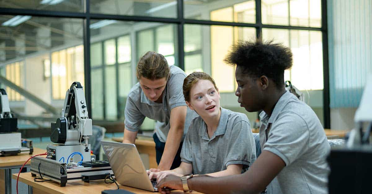 Three students wearing gray shirts collaborate at a desk with a laptop and a robotic arm, exploring AI technology in a classroom or lab setting.