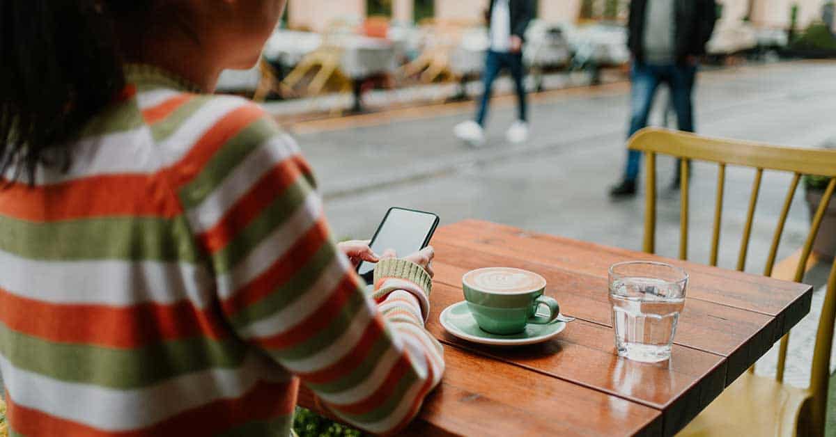 Person in a striped sweater sits at an outdoor café table with a coffee, a glass of water, and a smartphone, looking at the phone. Street and people are visible in the background—a reminder of the importance of staying secure when using public Wi-Fi in bustling shared spaces.