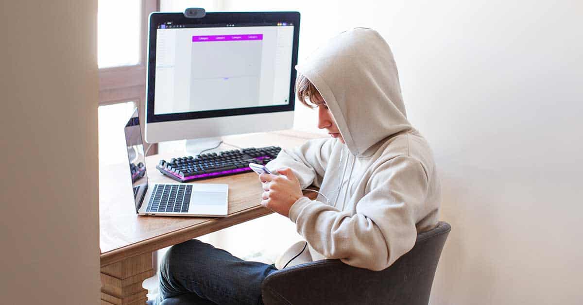 A person in a hoodie sits at a desk using a smartphone, with a laptop and a large desktop monitor in front of them, possibly involved in investigations related to online threats like sextortion.
