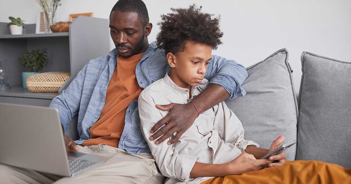 An adult and a child sit on a couch. The adult uses a laptop while the child looks at a smartphone, highlighting the importance of online safety and awareness of sextortion risks for young users.