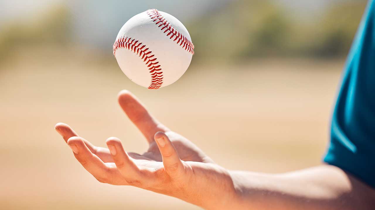 A person tosses a baseball into the air with their hand, outdoors in a blurred, sunlit setting during a Dorm trip after watching a Diamond Back game.