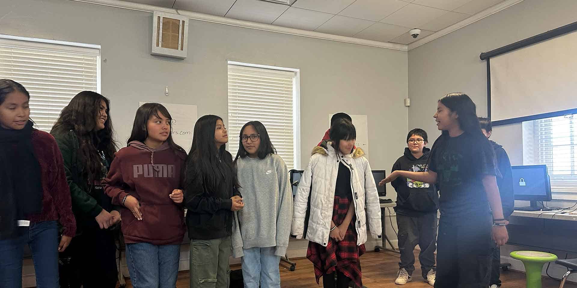 A group of students stands and talks together in a classroom with computers and windows in the background.