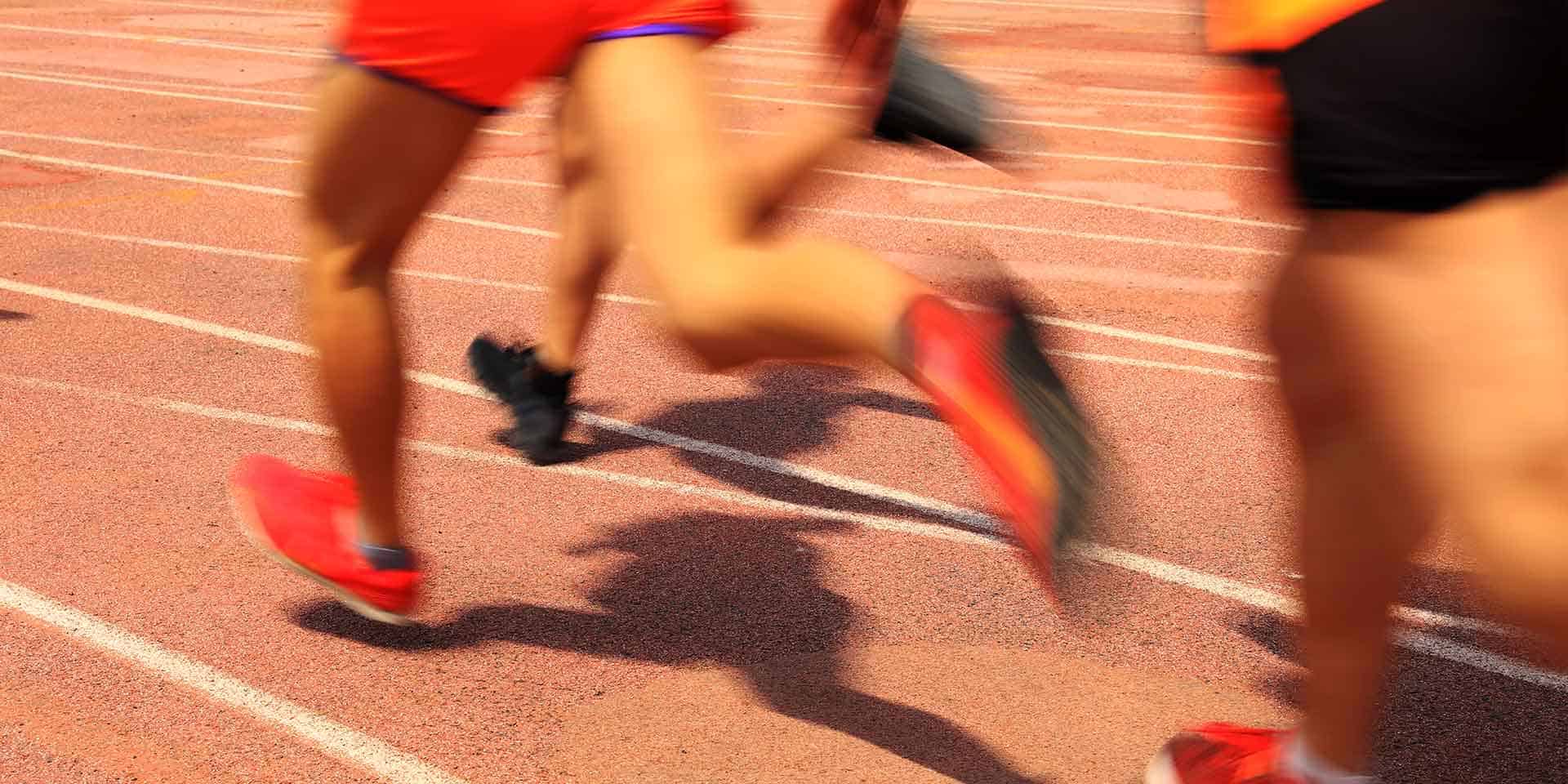 Three runners in motion on an outdoor track, with legs and shoes visible, captured in a blurred, dynamic action shot.