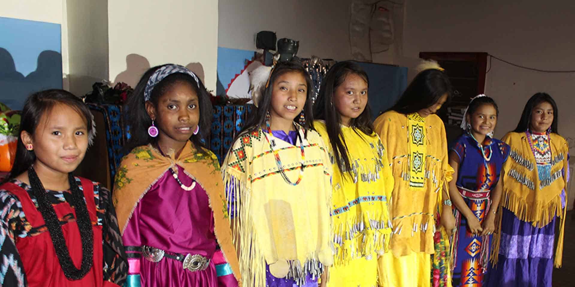 A group of girls in colorful traditional Native American dresses and accessories stand in a line indoors, facing the camera.