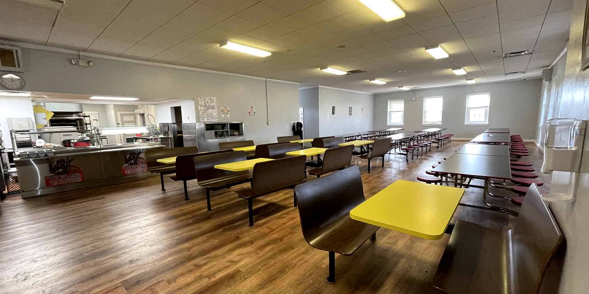 A cafeteria with yellow and brown tables, brown benches, a food serving counter on the left, and overhead fluorescent lighting. The room is empty.