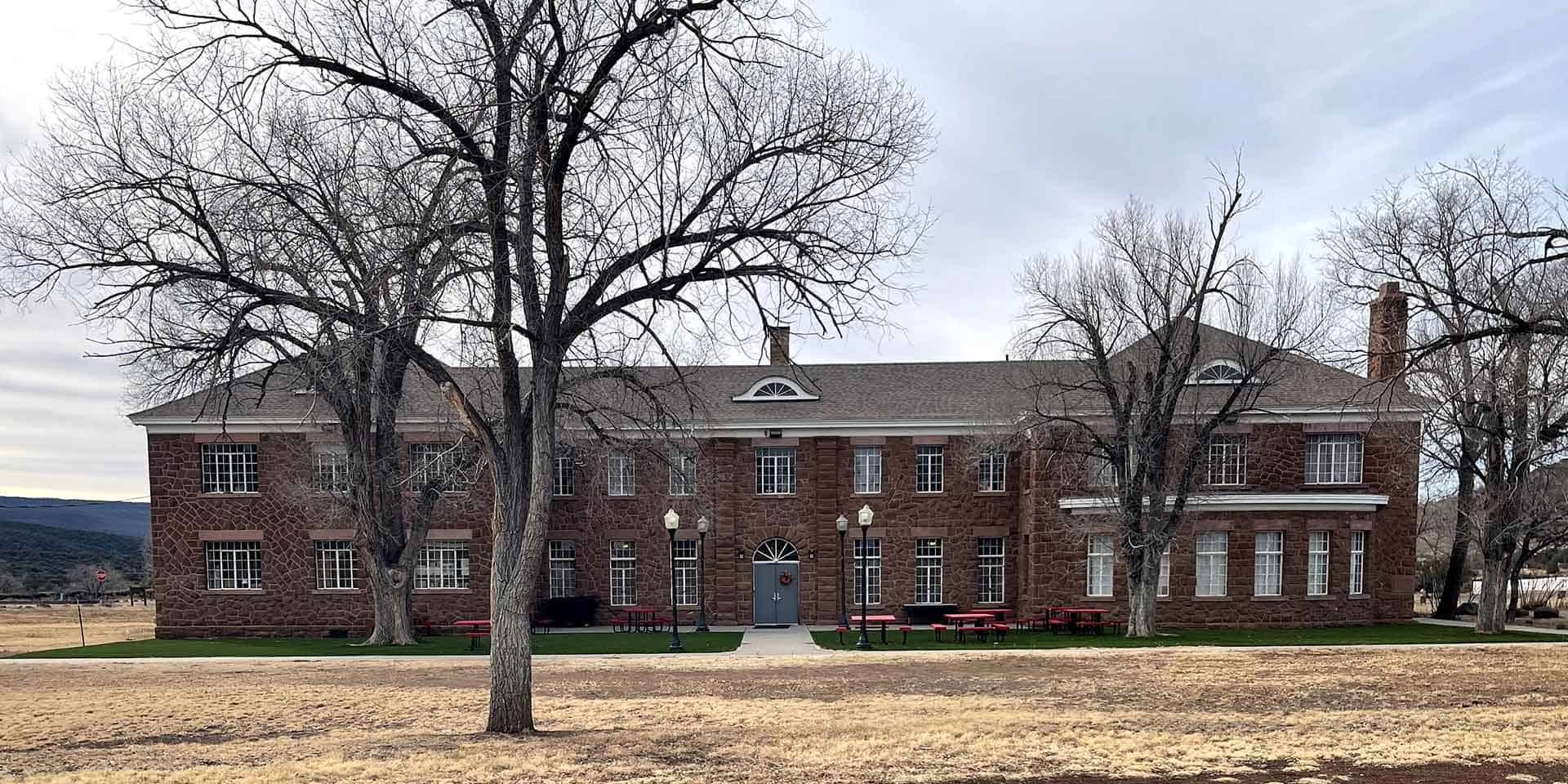 A two-story brick building with multiple windows, a central entrance, and leafless trees in front, set on a grassy area with red benches and lamp posts.