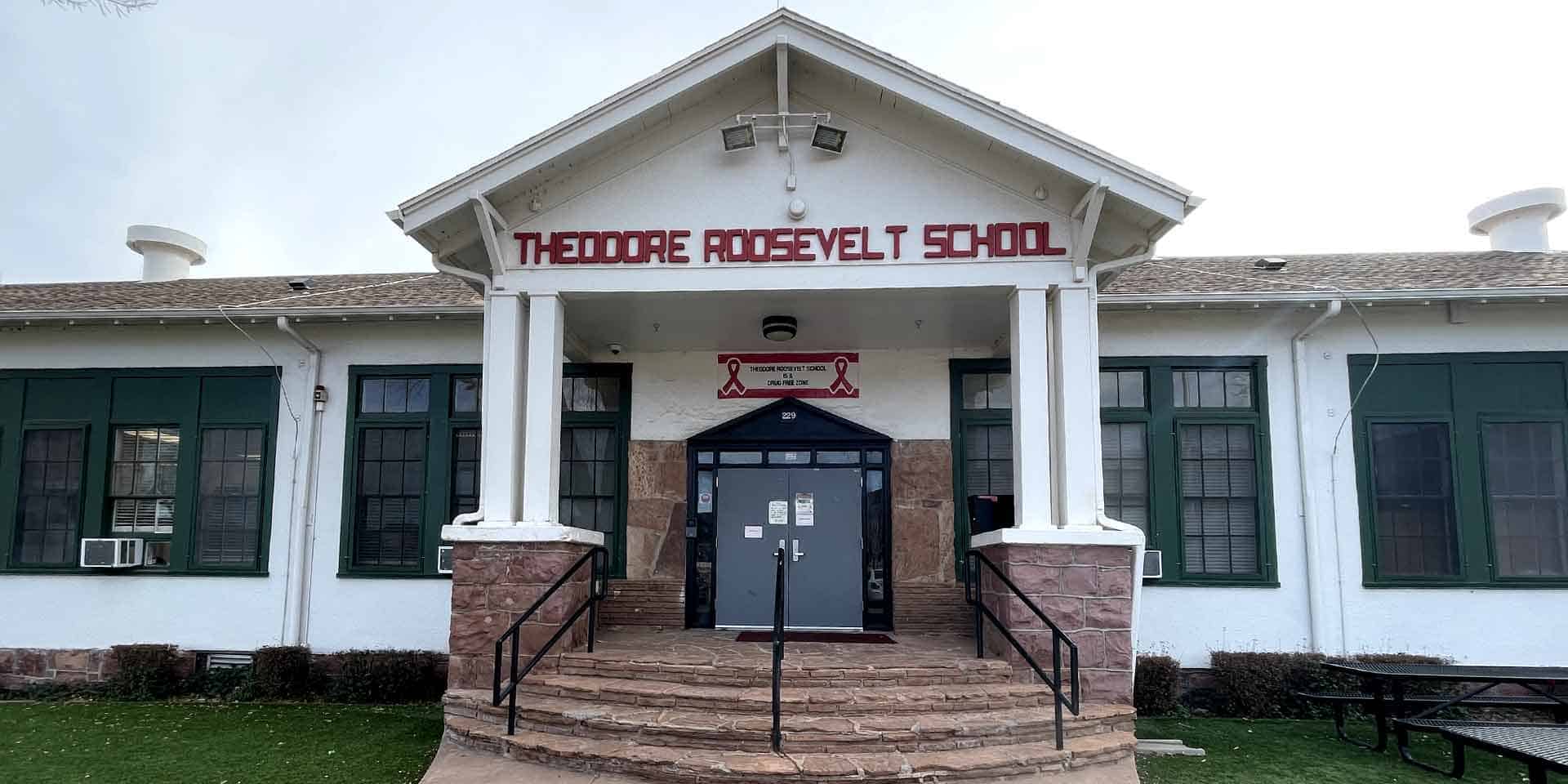Front entrance of Theodore Roosevelt School, a white building with green window frames, double doors, and a sign above the doorway.