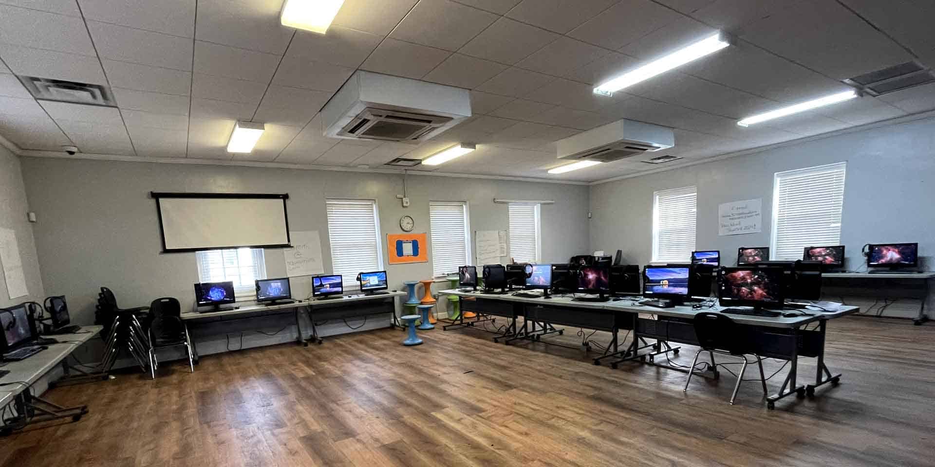 A classroom with multiple computers on tables along the walls, empty chairs, wood flooring, a projector screen, and fluorescent ceiling lights.