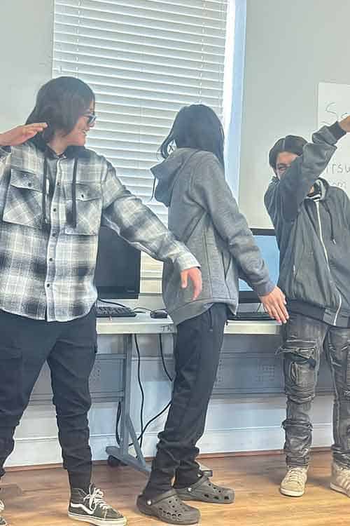 Three people stand indoors in front of computers, each posing with one arm bent and the other extended, appearing to mimic the letter "D" with their bodies.