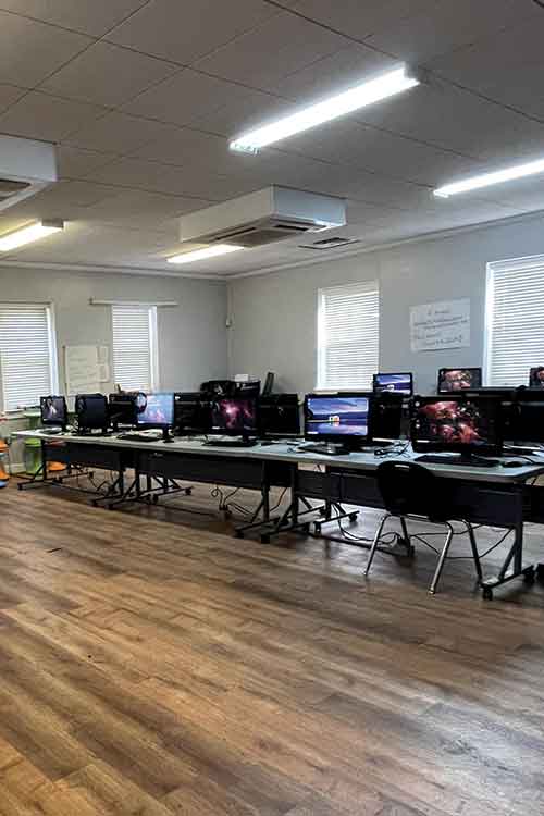 A classroom with multiple computer workstations on long tables, empty chairs, and wooden flooring, illuminated by ceiling lights and natural light from windows.