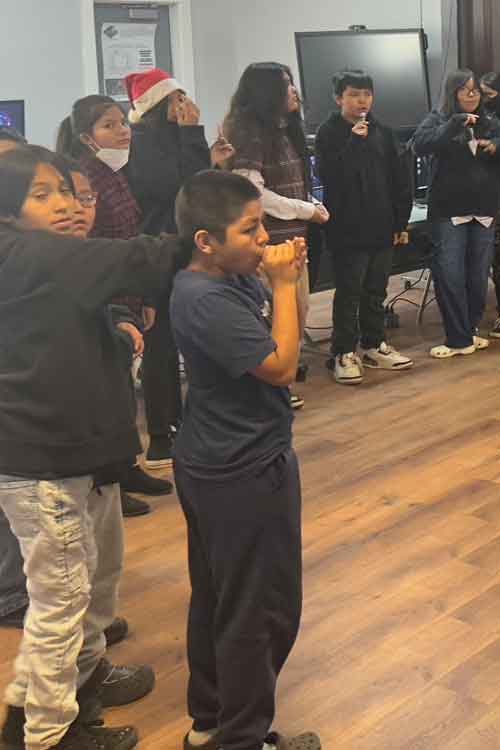 A group of children stands indoors, some holding their hands near their mouths or faces, with one child in the foreground and a Christmas hat visible on one person in the back.