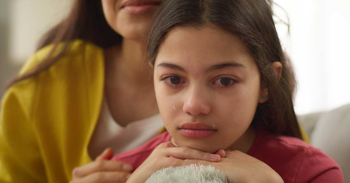 A young girl with tears on her face rests her chin on her hands, while an adult sits beside her, offering comfort after she confides about facing sextortion.