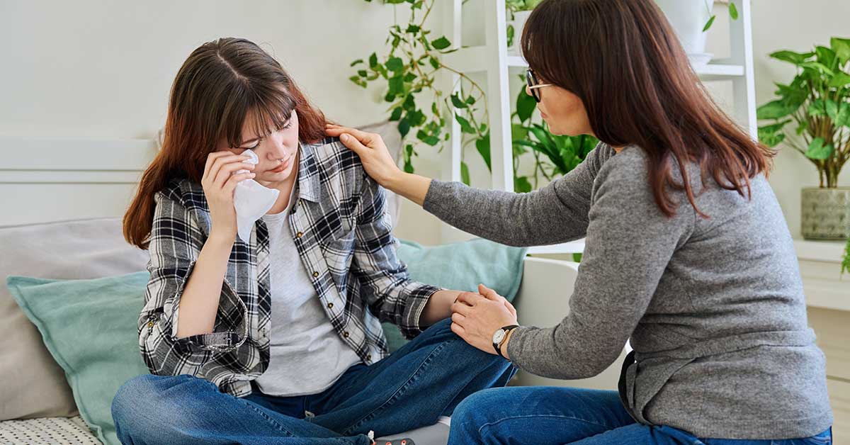 A woman comforts a young person who is sitting on a couch, crying and wiping their face with a tissue after experiencing sextortion.