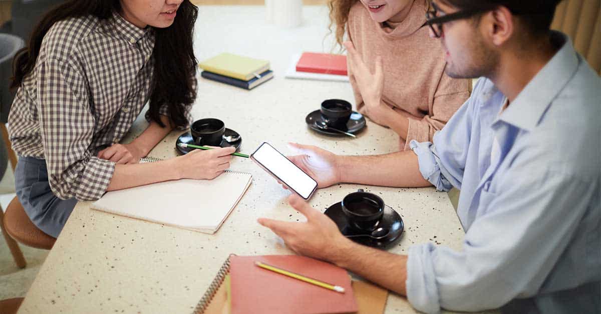 Three people sit at a table with notebooks, pencils, and coffee cups, having a discussion about AI while one person gestures and another holds a smartphone.