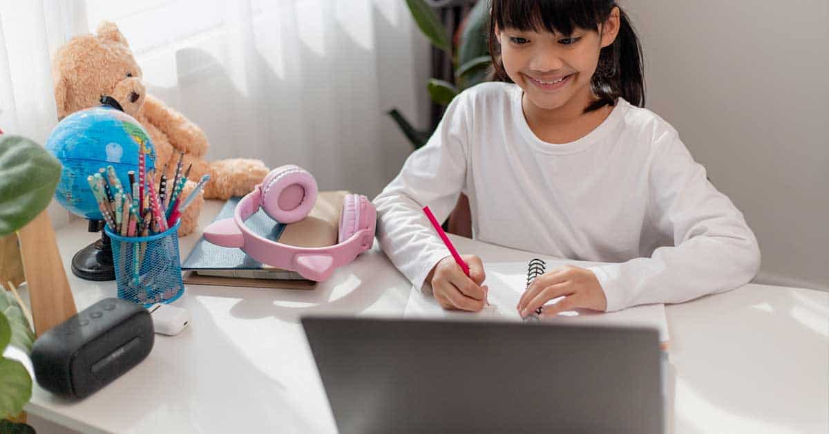 A girl in a white shirt sits at a desk, focused on her homework as she writes in a notebook and looks at her laptop. Headphones, a globe, and a teddy bear are also on the desk.