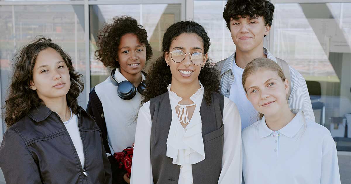 Five teenagers stand together outside a building, looking at the camera and smiling. Some wear casual clothes, one has headphones around their neck, and they all share an interest in AI literacy.