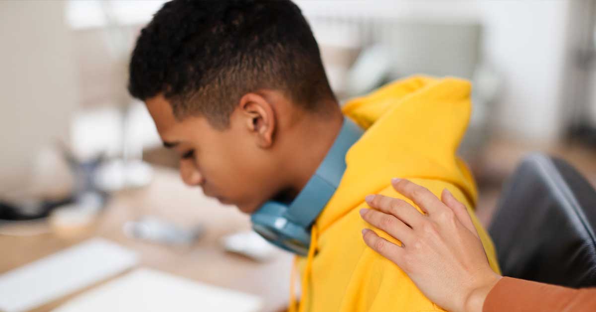A person wearing a yellow hoodie and headphones sits with a lowered head, possibly feeling the weight of sextortion, while another person's hand rests on their shoulder in a comforting gesture.
