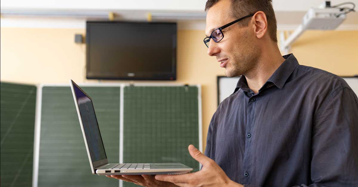 A man wearing glasses stands indoors holding an open laptop, with a TV and green boards in the background, possibly working on an AI project.