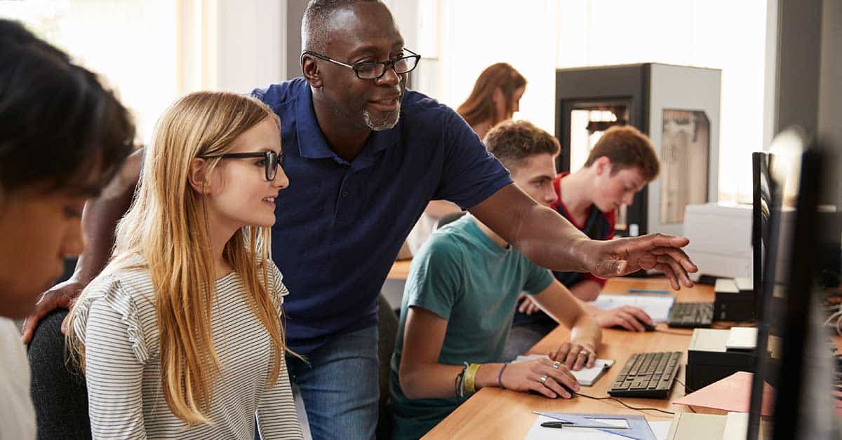 A teacher helps a student at a computer in a classroom, guiding them through a cybersecurity lesson while other students work at their computers in the background.