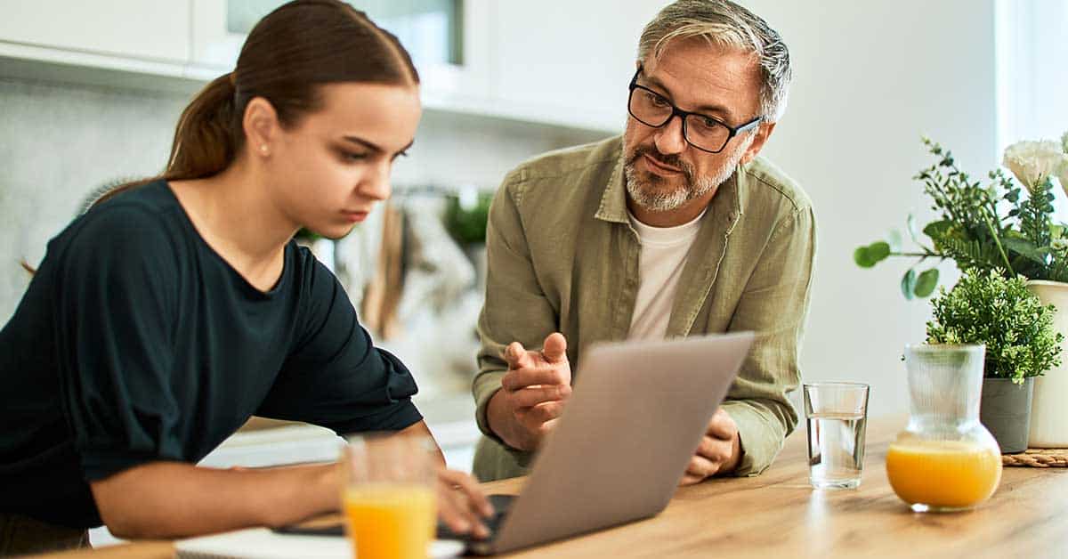 An older man gestures while talking to a young woman working on a laptop at a kitchen table, as drinks and plants sit nearby—an intense discussion that hints at blackmail.