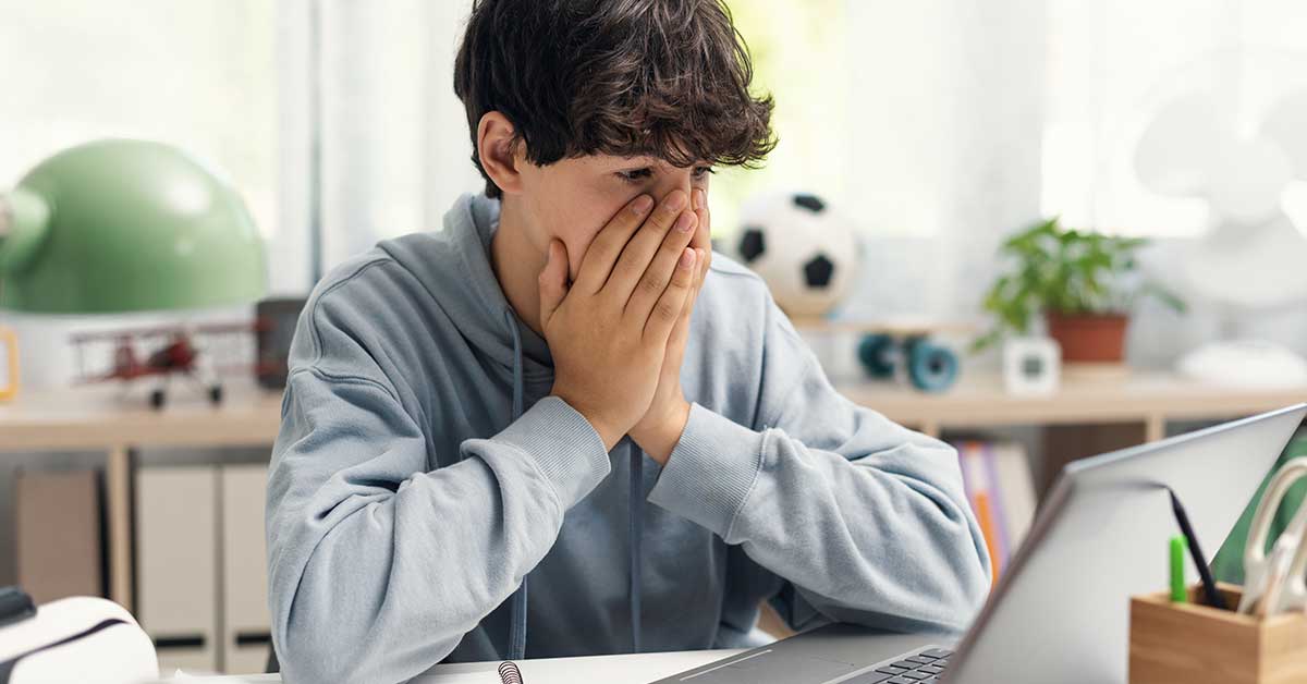 A young person in a hoodie sits at a desk, hands covering their mouth, looking at a laptop with concern—highlighting how sextortion is a hidden crime affecting many boys and young men.