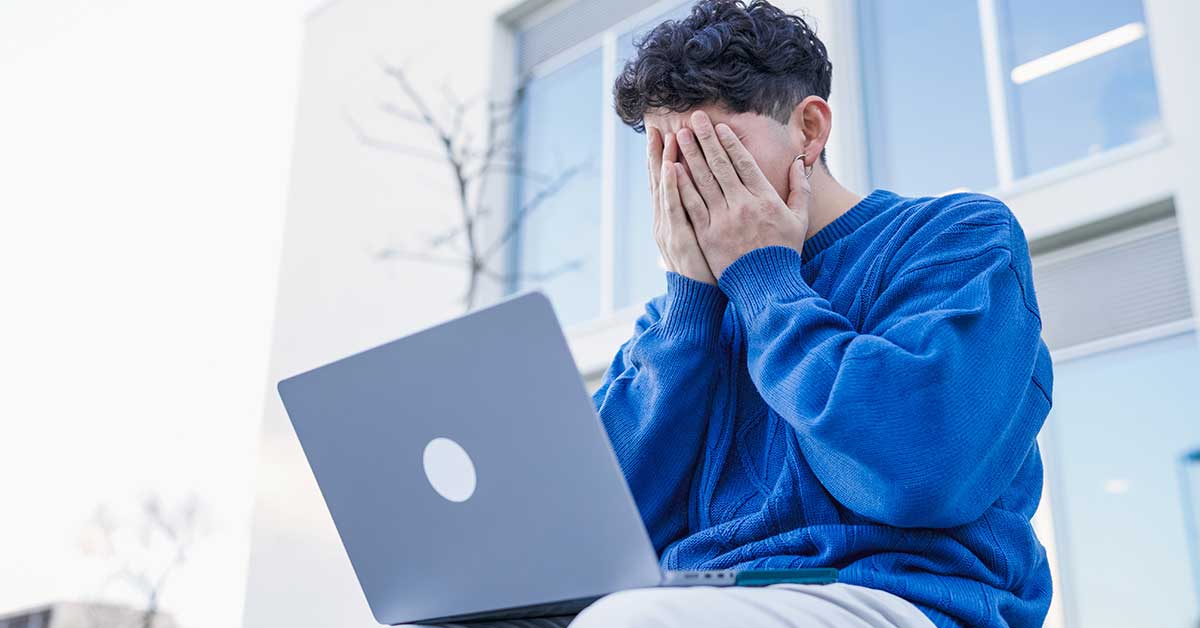 A person in a blue sweater sits outside with a laptop, covering their face with their hands in apparent frustration or distress—possibly worried about sextortion.