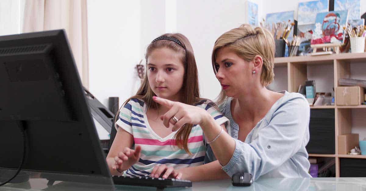 An adult woman sits beside a young girl at a desk, pointing at a computer screen while the girl types on the keyboard, as they discuss how to stay safe online and protect against dangers like sextortion.