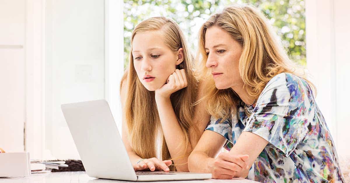 Two women sit at a table in a bright indoor setting, looking intently at a laptop screen as they discuss concerns about online sextortion.