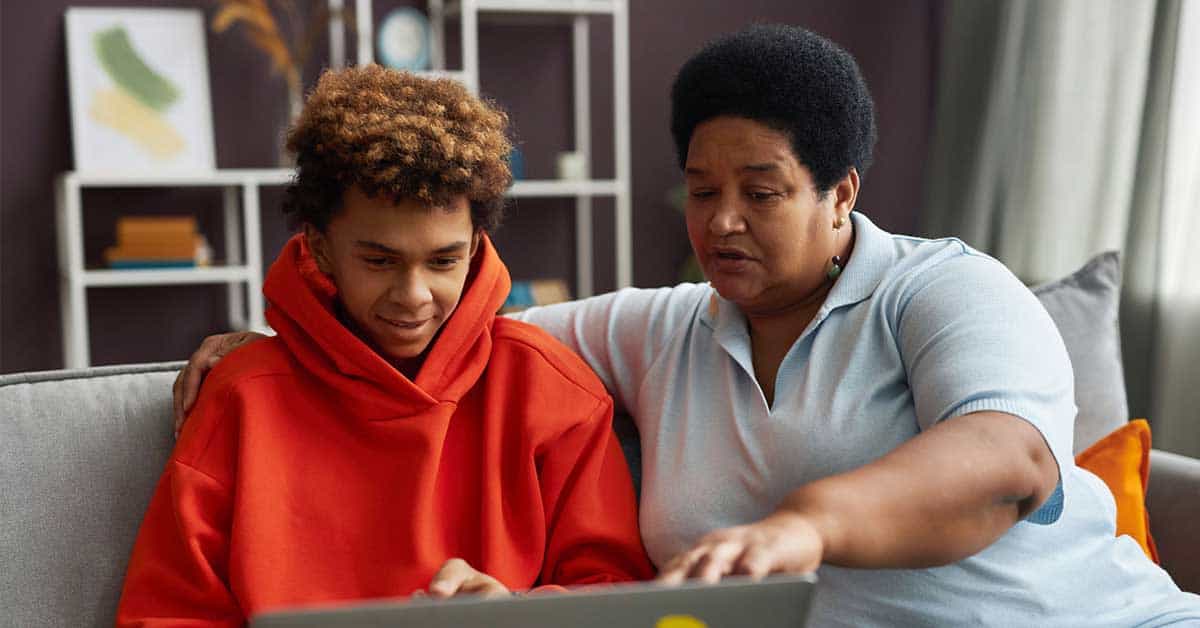 An adult and a young person sit on a couch, looking at a laptop together. The adult is pointing at the screen while discussing online safety, warning the teen about threats like sextortion.