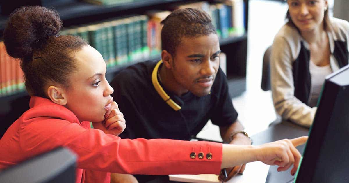 Three people sit at a desk with a computer in a library; one person points at the screen, encouraging critical thinking as the others watch attentively.